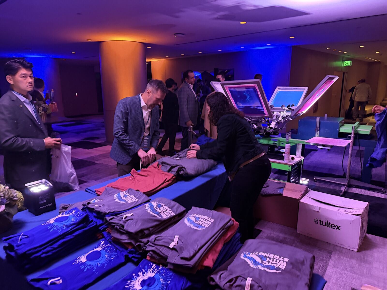 Guests selecting garments at a live personalization table during an evening corporate gala with blue uplighting