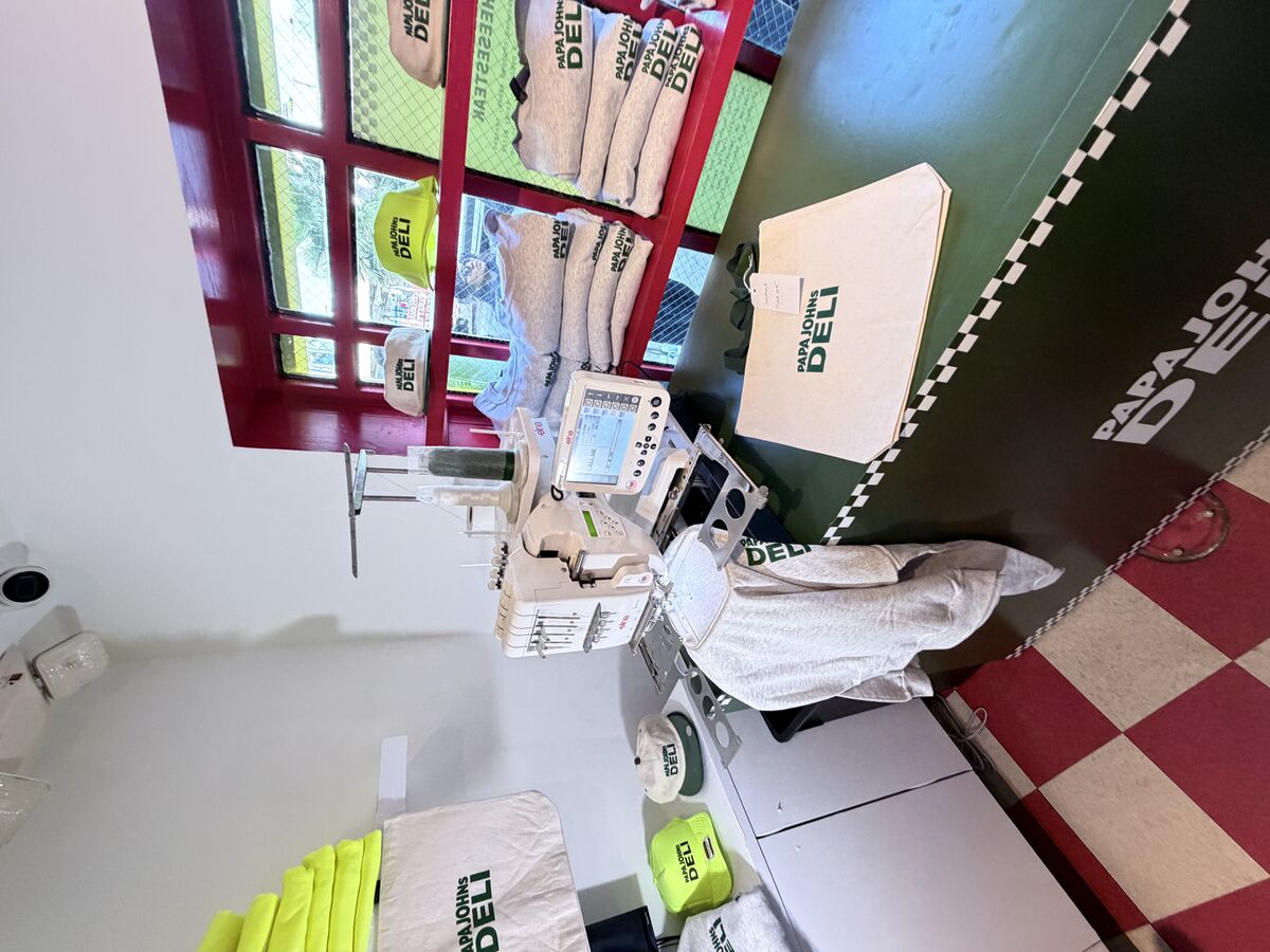 Embroidery machine stitching personalized pieces at a branded pop-up with finished caps, totes, and sweatshirts on display